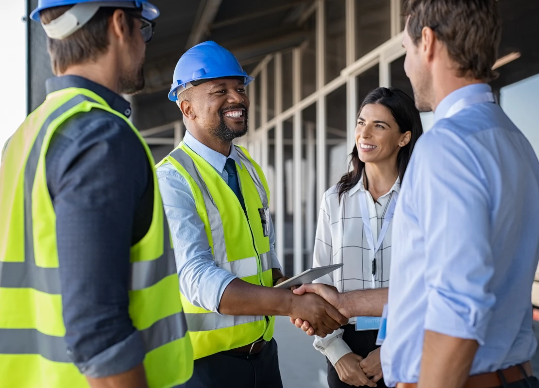 Smiling engineer shaking hands at construction site with happy architect. Handshake between cheerful african construction manager with businessman at bulding site. Team of workers with architects and contractor conclude an agreement with safety uniform.