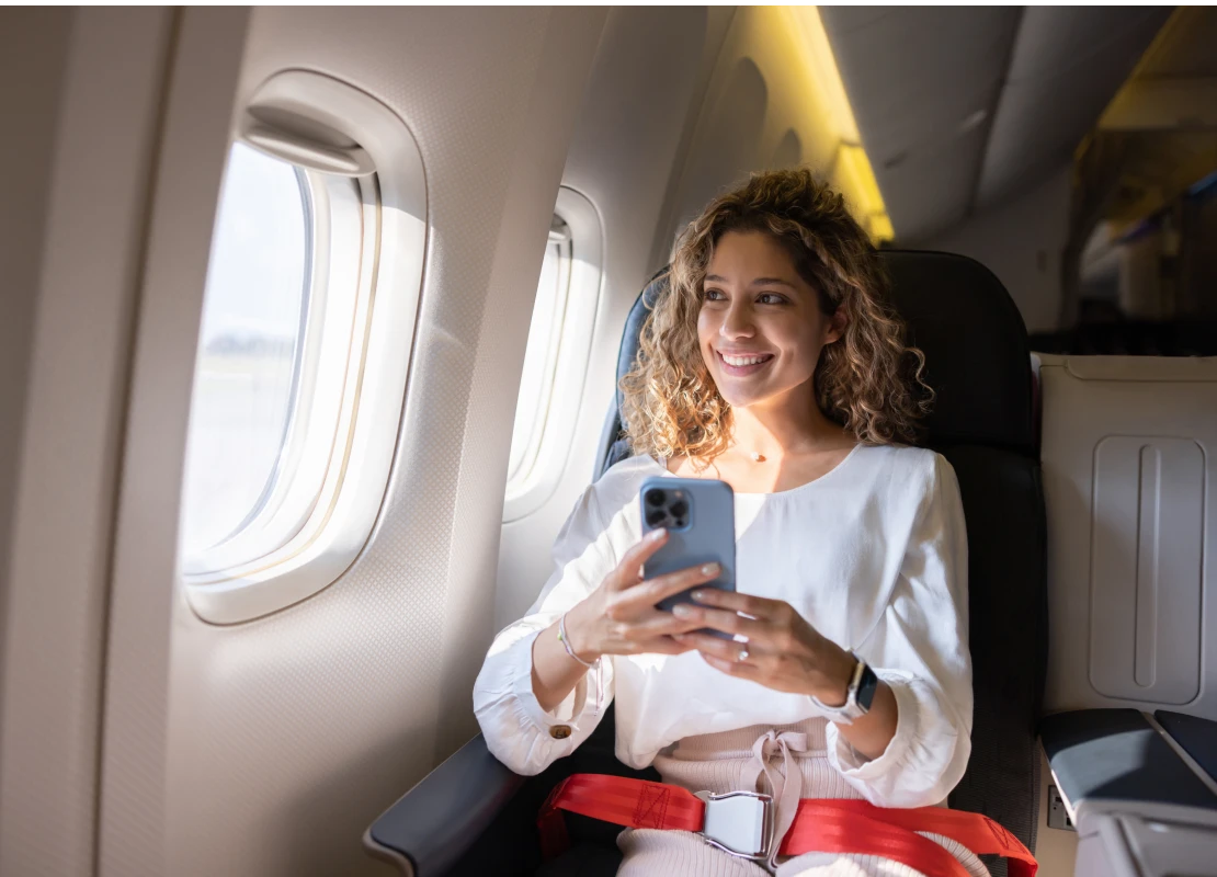 Latin American female traveler using her cell phone in an airplane and looking through the window